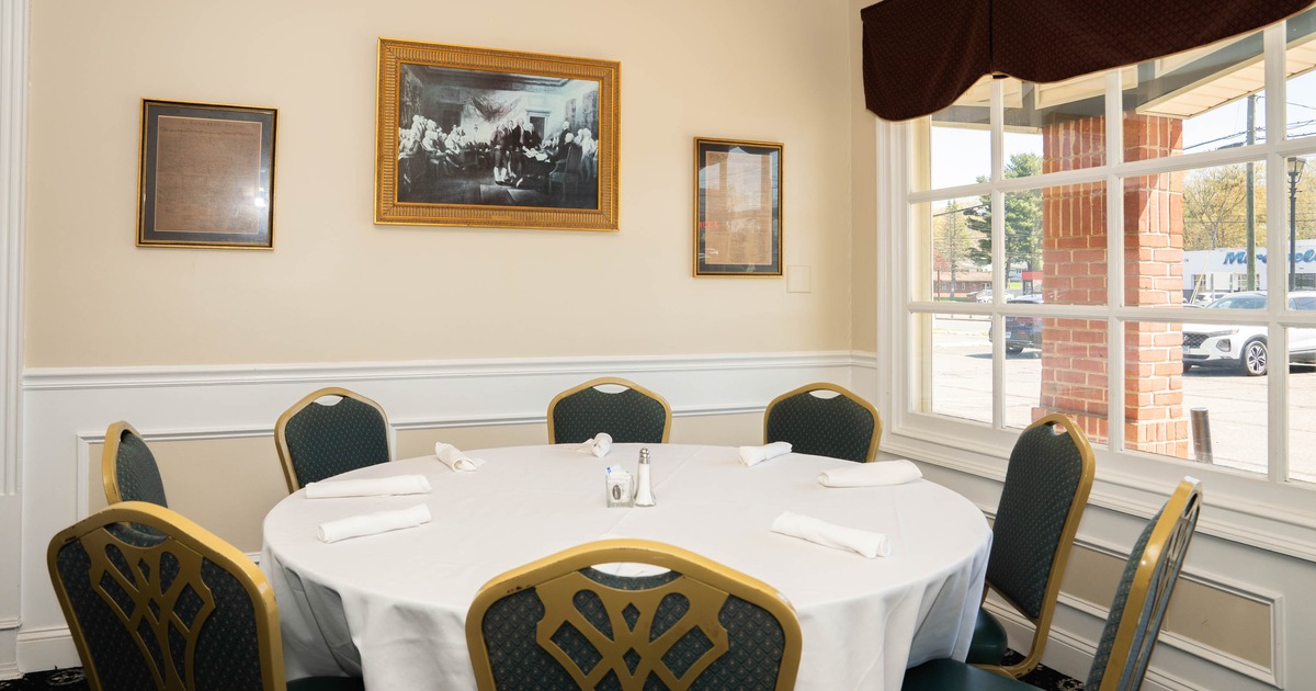 A round dining table with six chairs in a room with framed items on the wall and a window showing an outside view.