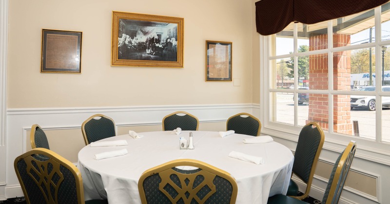 A round dining table with six chairs in a room with framed items on the wall and a window showing an outside view.