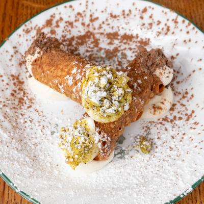 Cannoli with cream filling, pistachios, cocoa dusting, and powdered sugar on a plate.
