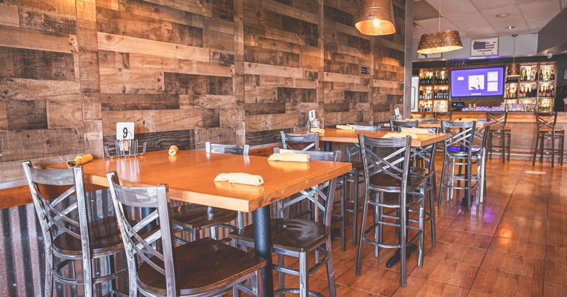 Interior, high wooden top seating tables with stools, wood coated wall, bar in the back