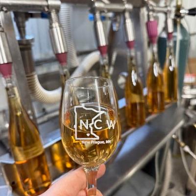 Hand holding a glass of white wine, featuring a NC logo, in front of a wine bottling machine.