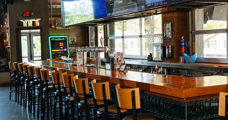 A modern bar with a long wooden counter, black stools and large TVs displaying sports.