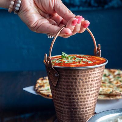 Hand holding a copper pail of red curry, garnished with cilantro, alongside naan and rice.
