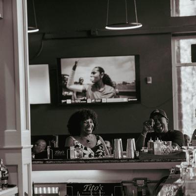 Interior, two people sitting at a bar, laughing warmly, behind them, a TV shows a sports scene.