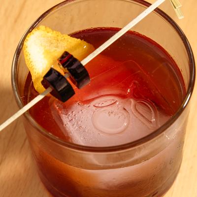 Red cocktail with a large ice cube and citrus garnish.