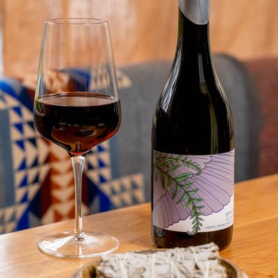 A glass and a bottle of red wine beside a sage smudge stick on a table.