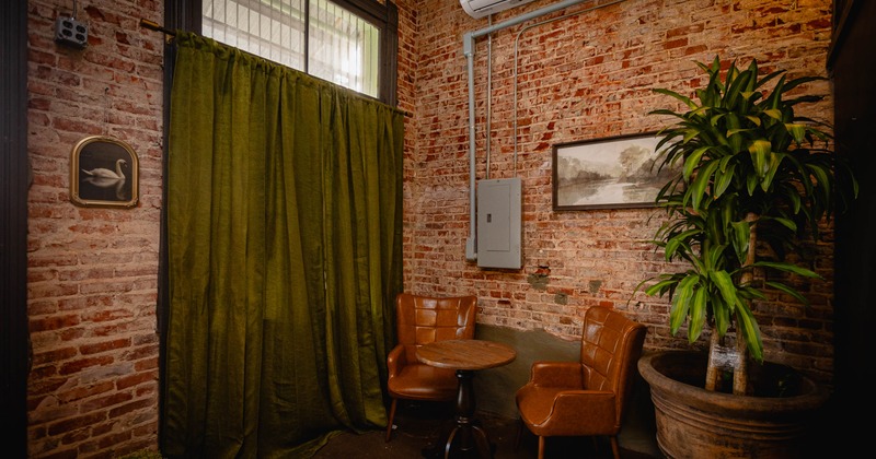 Two brown leather chairs, a wooden table, green curtains, exposed brick walls and a potted plant