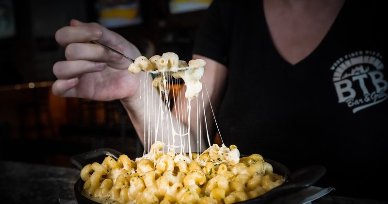 A person lifts a forkful of creamy macaroni and cheese from a skillet, with cheese stretching