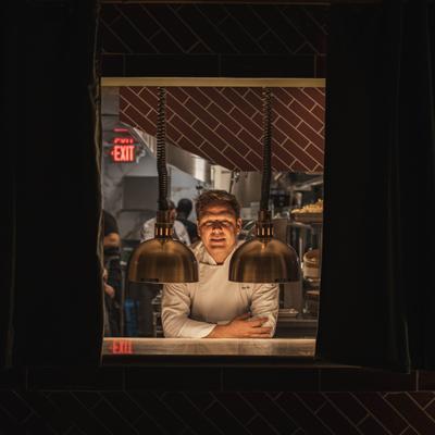Chef standing at the kitchen pass under heat lamps, viewed through the service window.