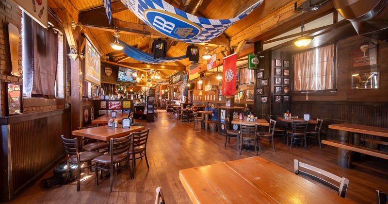 Interior with wooden tables, chairs, exposed beams, hanging flags, and a lit bar area in the background