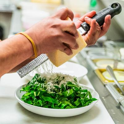 Hands grating cheese over a bowl of fresh greens.