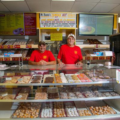 Two smiling owners stand behind a glass display case filled with dozens of donuts and pastries.