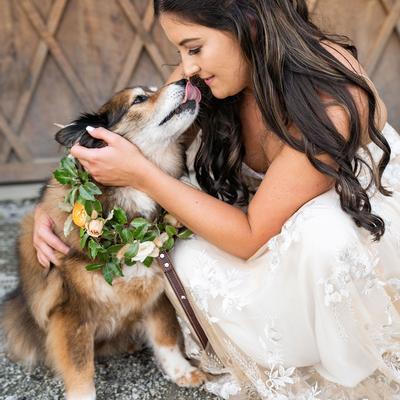 A bride in a white lace dress lovingly embraces a dog wearing a floral collar