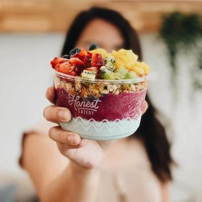 A person holds a colorful acai bowl topped with fruits and granola, with Honest Eatery on the bowl.