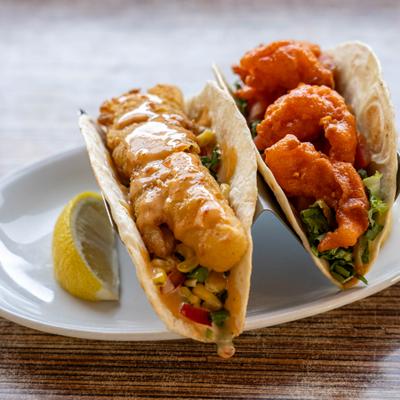 Shrimp taco and fish taco served on a white plate with a lemon wedge.