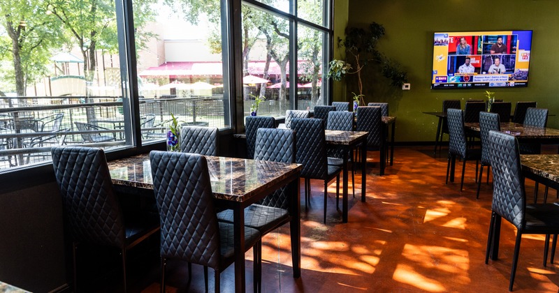 Empty restaurant dining room featuring large windows, dark quilted chairs, and a television