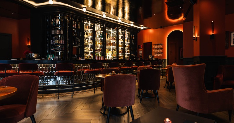 A warmly lit bar with red velvet chairs, wooden herringbone flooring, and bottles on display behind the counter