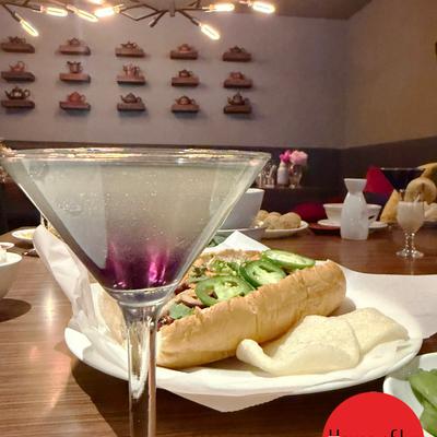A cocktail in a martini glass, sits beside a plated Banh mi sandwich.