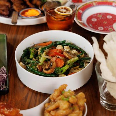 A bowl of sauteed mixed vegetables sits on a wooden table, surrounded by food and drinks.