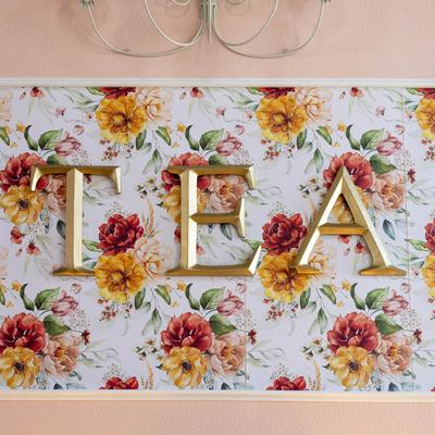 Gold letters TEA mounted on a floral-patterned wall panel.
