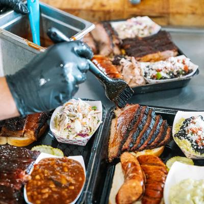 A gloved hand brushes sauce onto a tray of barbecue meats.