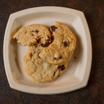 Two chocolate chip cookies are on a beige square plate, set on a dark surface.