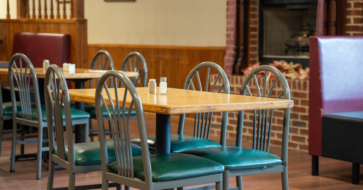 A cozy interior with wooden tables, green chairs and a brick fireplace in the background.