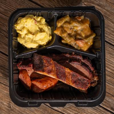 Takeout tray with ribs, sausage, potato salad, and stewed vegetables.