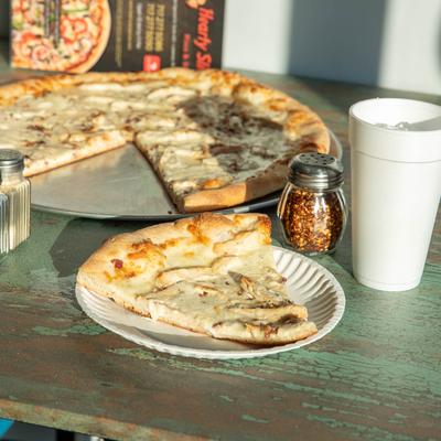 A slice of pizza on a paper plate, with a whole pizza, condiments, and a drink.
