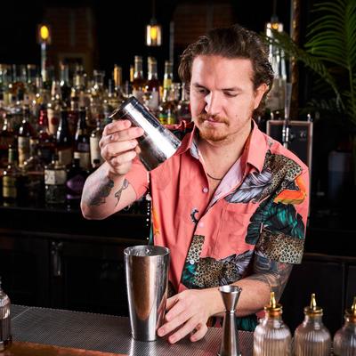 A bartender pours liquid into a metal shaker behind a bar lined with bitters bottles.