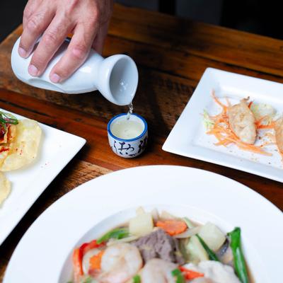 Hand pouring sake into a small cup on a wooden table surrounded by plates of Asian cuisin.