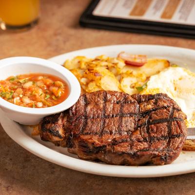 Grilled ribeye steak served with eggs, potatoes, and salsa on a plate.