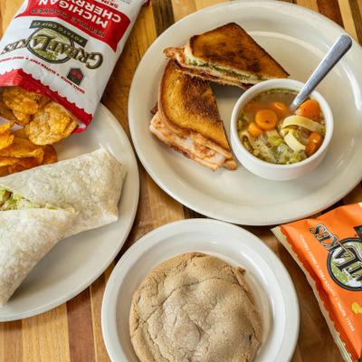 Turkey pesto melt with a bowl of soup, beside a bag of chips, a halved wrap, and a cookie.