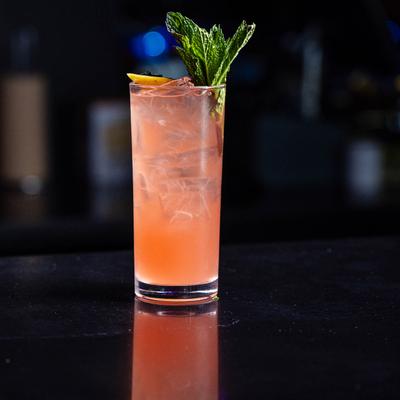 A tall glass of pink cocktail on a dark surface, garnished with a lemon slice and fresh mint leaves.