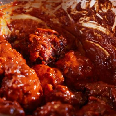 Close-up view of glossy, dark red, saucy chicken pieces being tossed in a metal mixing bowl.