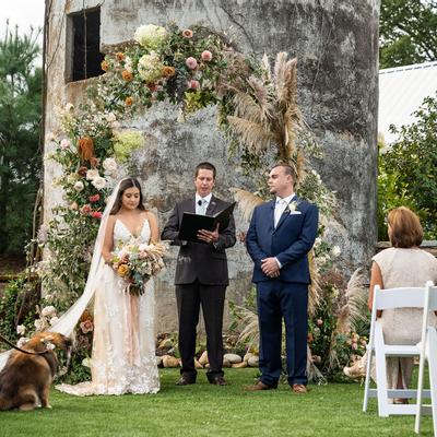 A bride and groom stand before a floral arch with an officiant at an outdoor wedding