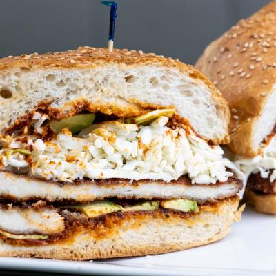 Sandwich with sesame seed bread, avocado, fried meat, shredded cabbage, sauce.