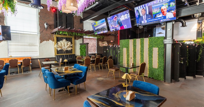 A modern restaurant interior with blue velvet chairs, black tables, hanging foliage, and multiple televisions