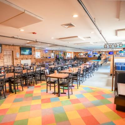 Restaurant interior with colorful carpet, wooden tables, black chairs, and a bar in the background..