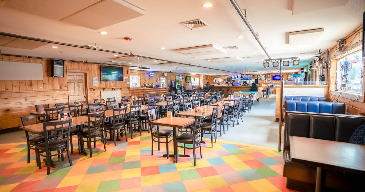 Restaurant interior with colorful carpet, wooden tables, black chairs, and a bar in the background.