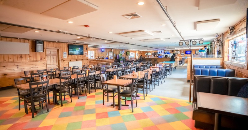 Restaurant interior with colorful carpet, wooden tables, black chairs, and a bar in the background.