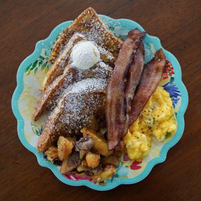 Breakfast plate with French toast, scrambled eggs, crispy bacon, and smashed potatoes.