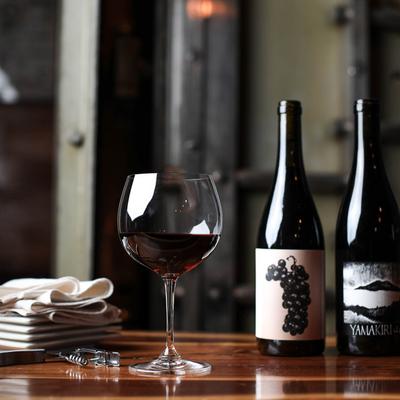 Glass of red with alongside two bottles, corkscrew and wine cloth.