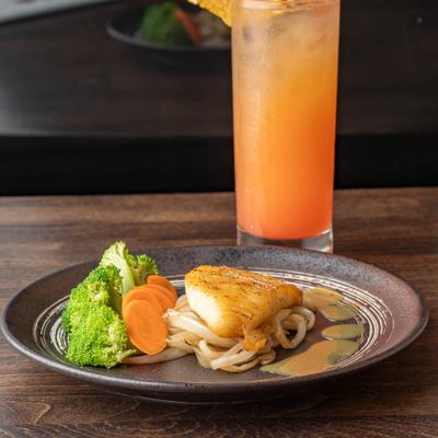 Seared fish with noodles, vegetables, and orange cocktail on a wooden table.