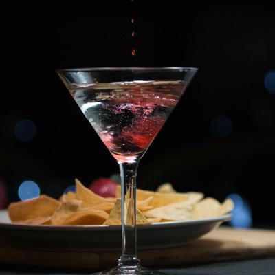 A martini glass sits on a table, with a plate of chips against a dark background.