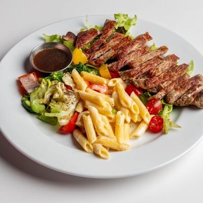 A plate holds sliced steak, pasta with a creamy sauce, mixed vegetables, dipping sauce.