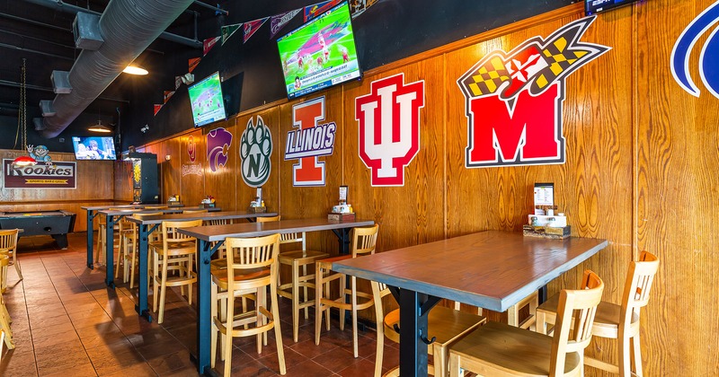 Interior, lined up tables and bar chairs by a wall with TVs and sports logo stickers