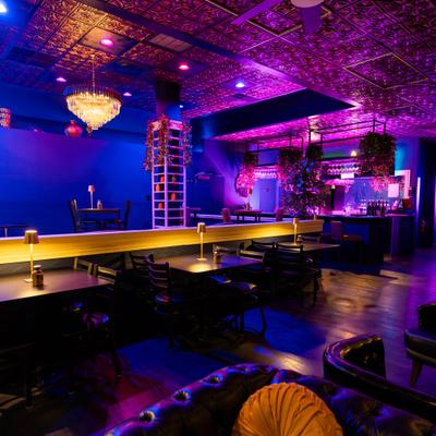 Bar interior with vibrant blue and purple lighting, tufted seating, and a chandelier.
