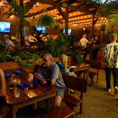 Patrons sitting at tables in a dimly lit outdoor patio bar with string lights and tropical plants.