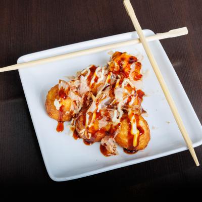 Takoyaki drizzled with sauces and bonito flakes on a white plate with chopsticks.